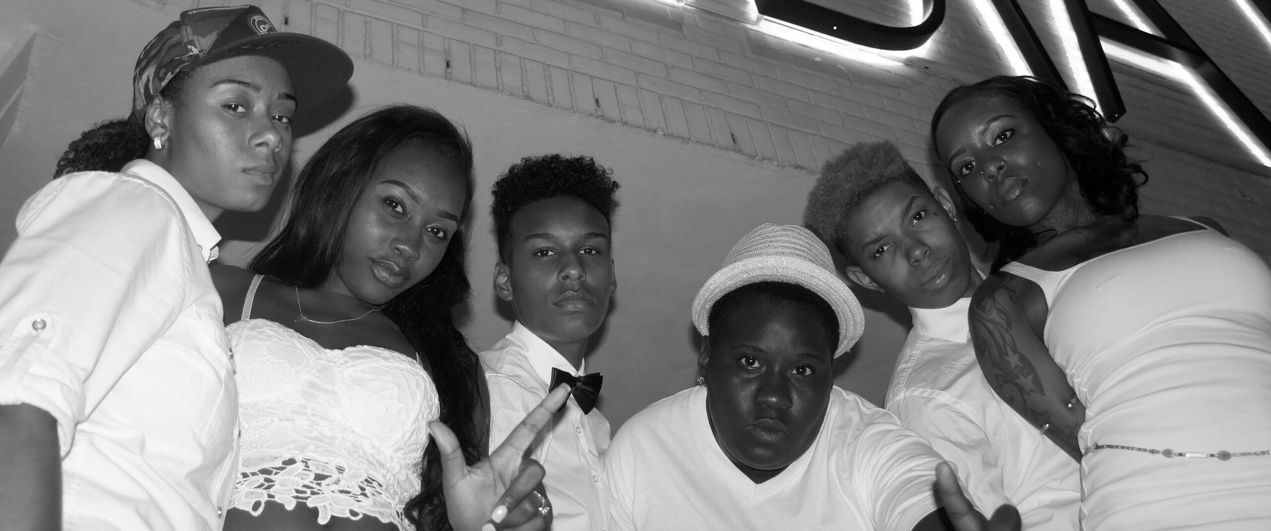 Black-and-white photo of six masculine- and femme-presenting Black and Brown queer individuals posing confidently beneath bold signage on a city street at night. They wear crisp white attire ranging from lace crop tops and fitted bow ties to button-downs and hats. Each gaze meets the camera with unapologetic energy, showcasing individual style, queer identity, and community unity.
