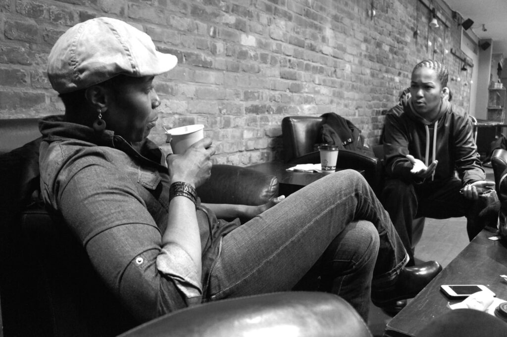 Alt Text (SEO + Accessibility Optimized): Black-and-white photo of two masculine-presenting Black individuals in relaxed conversation inside a cozy café with exposed brick walls. One sits back with a coffee cup in hand, dressed in a cap, denim shirt, and earrings. The other leans forward intently, wearing braids and a hoodie, gesturing expressively. Phones, jackets, and another coffee cup sit casually on nearby tables, evoking a laid-back yet engaged atmosphere.