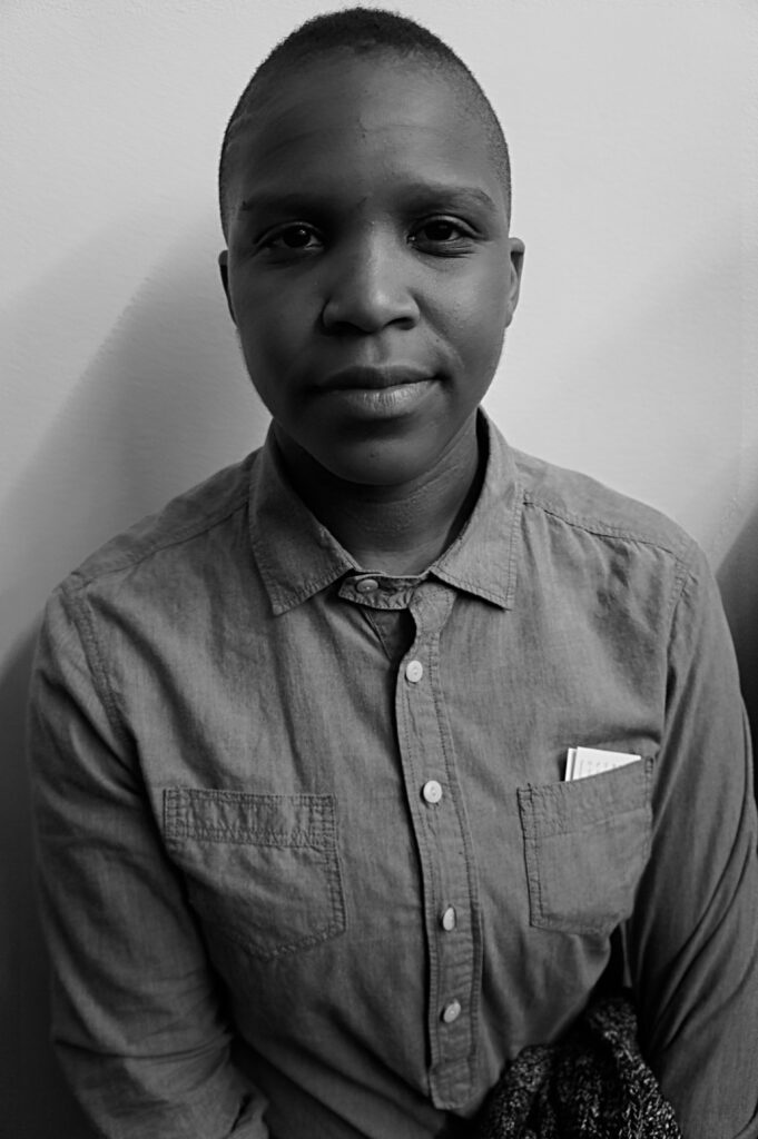 Black-and-white portrait of a dark-skinned, masculine-presenting individual wearing a button-up chambray shirt. They sit calmly against a plain white wall, making direct eye contact with the camera. A small paper notebook peeks from their chest pocket, adding texture to the intimate, minimalist setting.