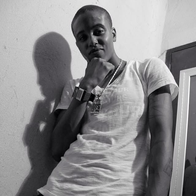 Close-up black-and-white portrait of a masculine-presenting Black individual with long locs and a white beanie. Their eyes look directly into the camera with calm intensity and quiet strength, offering an unguarded moment of presence and self-awareness.